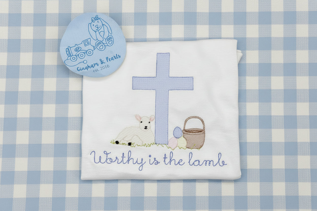 white baby shirt with an illustration of a lamb, a cross, and the text 'Worthy is the lamb' on it, with a blue and brown design accent.