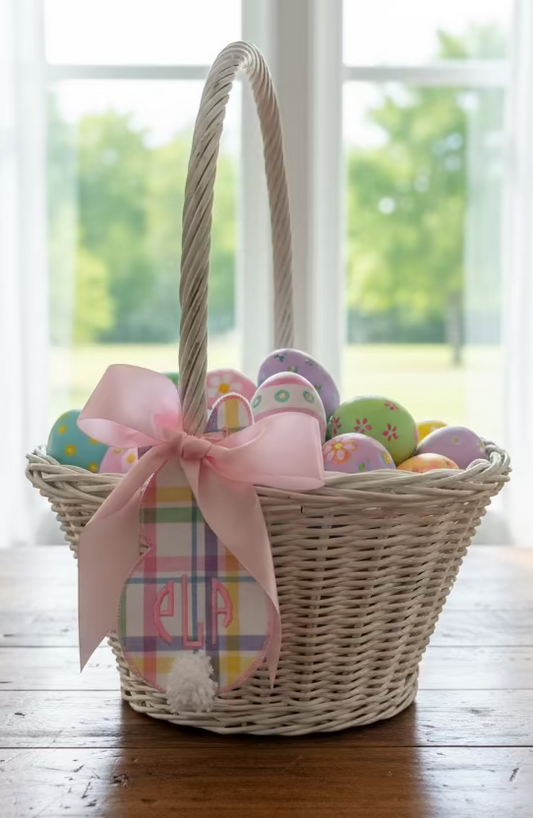 A wicker basket with a white handle and a pink ribbon bow, adorned with a blue and white plaid tag with a monogram on it.